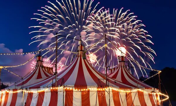A red and white striped circus tent illuminated by fireworks and string lights at dusk.
