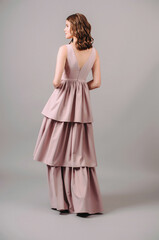 Fototapeta premium Sensual portrait of brunette woman in powder evening dress walking in studio. Bridesmaid's fashion. Dust pink plunge neckline sleeveless maxi gown with tiered skirt. Summer feminine look, rear view