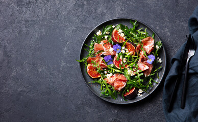 Arugula salad with fresh figs and prosciutto, jamon and edible flowers in a plate. Dark grey background. Copy space. Top view.
