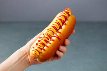 A hand holds a hot dog with ketchup and mustard. Grey background. Close up.