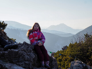 Naklejka premium Young Girl in Pink Winter Jacket Sitting on Mountain Rock with Panoramic Mountain View