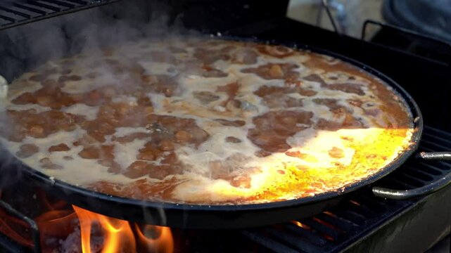Large pan with traditional spanish paella cooking over a wood fire on a grill, creating steam and delicious aromas
