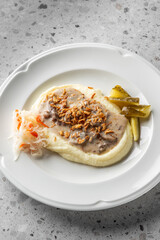 A plate of creamy mashed potatoes topped with savory gravy and crispy onions, accompanied by pickles and a side of shredded cabbage. The dish is presented elegantly on a textured surface.