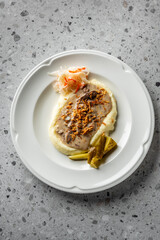 A plate of creamy mashed potatoes topped with savory gravy and crispy onions, accompanied by pickles and a side of shredded cabbage. The dish is presented elegantly on a textured surface.