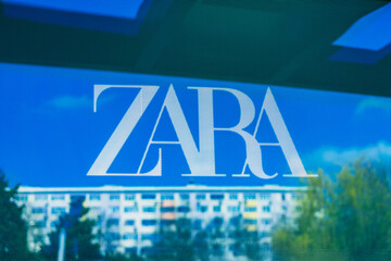 Fototapeta premium White ZARA logo on perforated glass, reflection in city street shop window in Burgas, Bulgaria, blurred urban buildings, sky in background. Fashion brand and cityscape in modern visual contrast
