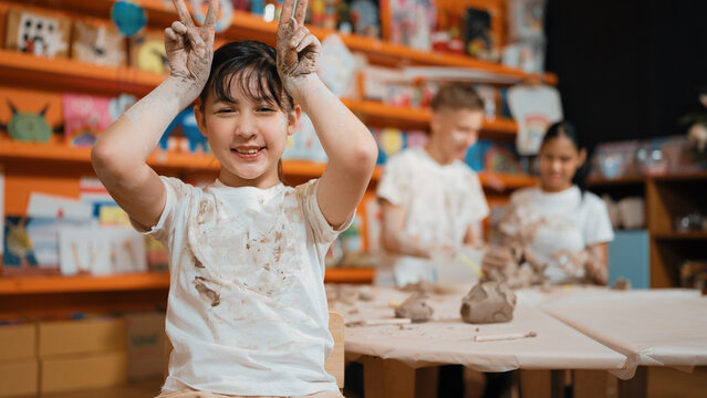 Asian girl pose at camera while diverse children modeling clay behind. Happy cute student wearing dirty shirt while looking at camera at workshop in art lesson. Blurring background. Edification.