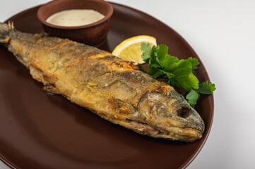 Crispy fried whole fish on a light background, on a brown plate with fresh parsley, a slice of lemon and a creamy sauce. A delicious seafood dish suitable for a restaurant menu or a cooking blog