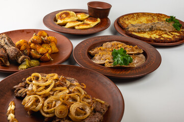 Traditional Eastern European cuisine served on rustic clay plates. A variety of homemade dishes including meat, potatoes, pancakes, dumplings and grilled vegetables, on a light background