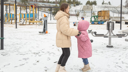 Young mother and a little girl in warm outerwear walk in the park in the children's playground in...