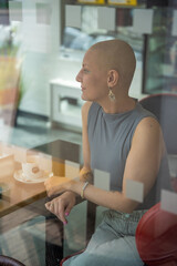 Fototapeta premium Young woman with cancer sitting inside a cafe, seen through the window, lost in thought. The concept highlights how people with serious illnesses continue to live actively and be part of society