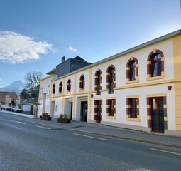 A two-story building with arched windows, yellow and red brick facade, and a modern extension on the roof. It is located along a street with yellow road markings and a sidewalk with planters.