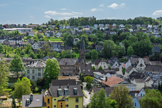 A scenic hillside view of Engelskirchen-R&uuml;nderoth, Germany, featuring the Evangelical Church at the center, surrounded by historic buildings, greenery, and the town square.

