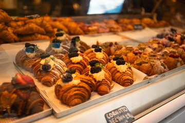 A variety of fresh, golden croissants and pastries are displayed in a bakery showcase. The croissants are topped with colorful fruits and cream, inviting customers to indulge in these delicious treats