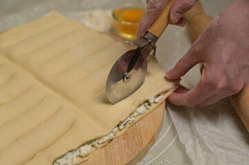 Close up square sheet of dough cut into strips with woman's hands on wooden board. Cutter, flour, egg on kitchen table. Home cooking baking puffs