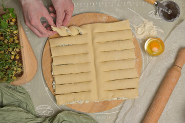 Top view woman's hands twists dough strips on wooden board. Cutter, egg, green towel, microgreens sprouts on kitchen table. Home cooking baking puffs