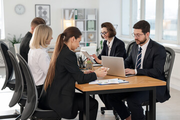 Coworkers with different devices working together at wooden table in office