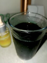A clear glass filled with a dark, chilled beverage with condensation on the surface. A small bottle with a dropper of yellow liquid sits beside it. The dim lighting adds a moody ambiance.