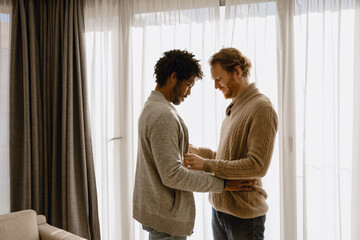 Fototapeta premium Gay couple, two men standing and holding hands next to each other while looking down, in the living room
