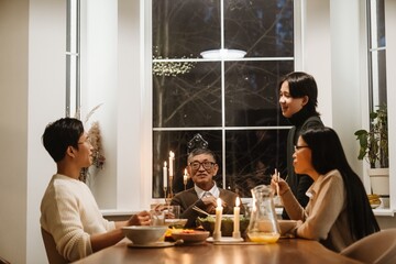 A grandson stands and talks while his father, grandfather, and grandmother sit at the table next to him, during dinner