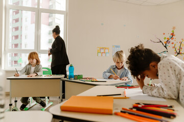 A group of three schoolchildren sit at desks and write while a teacher stands next to them looking out the window, at school, in the classroom