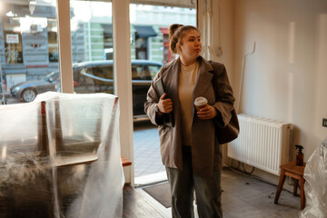 The woman owner is standing and holding a computer tablet and a cup while looking to the side,...