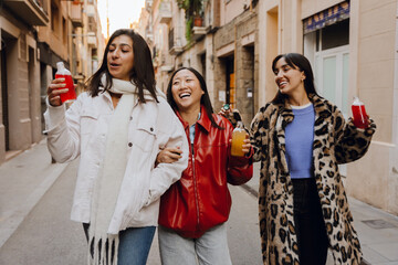 Obraz premium Group of three female friends walking and laughing while talking and holding bottles, outdoors, at a girls' party