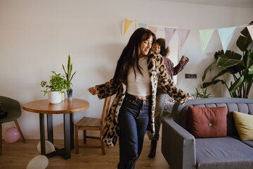 A woman runs next to a sofa while her female friend runs behind her, they are smiling, at a girls' party
