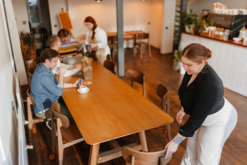 Obraz premium A female owner stands and holds a chair while a group of three customers sit at a table next to her and a female employee stands, in a cafe