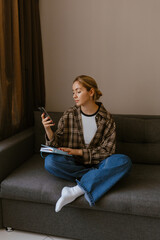 Obraz premium A woman sits on a sofa and holds a notebook while looking at her phone in front of her, in the living room, preparing for a trip