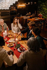 Family and guests sit at a table and watch as a man sitting between them cuts a pie during Christmas dinner