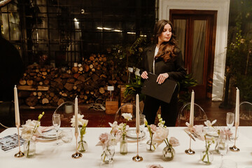 A woman organizer is standing and holding a clipboard while looking at the table next to her, in the venue, preparing for the wedding