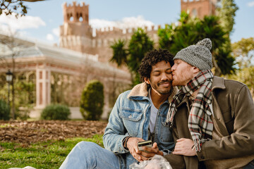 Obraz premium A man sits and kisses the cheek of a man sitting next to him and smiling while holding a phone, outdoors, on a date