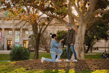 A man stands on one knee and puts a ring on the hand of a man standing across from him, on the street, on a date
