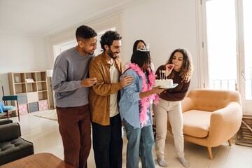 A group of five friends stand and laugh while one of them holds a cake and one of them blows a birthday pipe at a birthday party