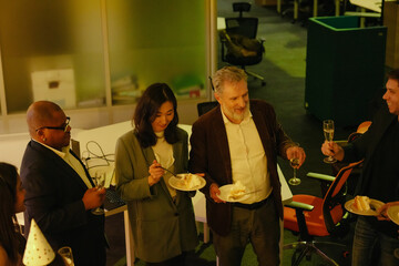A group of five employees stand and smile while holding glasses and plates with pieces of cake, in the hall, at a retirement party