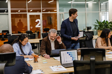 In a business meeting, a White man in his 60s works on a laptop while sitting at a long table surrounded by his colleagues of different ethnicities. Concept of an office work, diversity, inclusivity.
