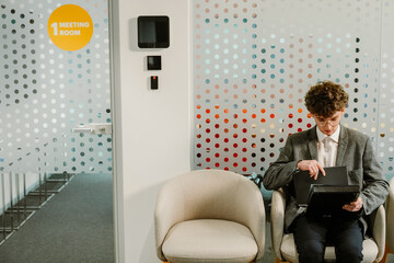 A young White man sits on a beige chair in a business premises against the white perforated wall and examines a black folder in his hands. Concept of waiting for an interview, preparing for a new job.