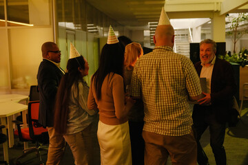 A White elder man and a multinational group of his co-workers wearing party hats, celebrating his...