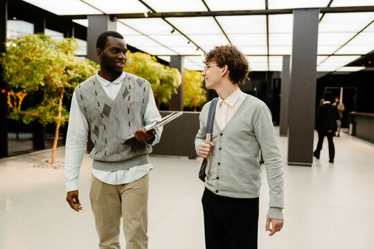A young black man and a white man walk side by side in the modern office lobby, casually talking and holding laptops and a bag. Business context with professional attire and bright space.