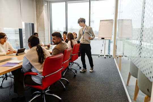 A White young man with glasses is presenting to a multinational group of people during his internship, while engaging in a collaborative discussion in a modern and clean office environment.