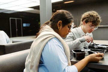 Obraz premium A young olive-skinned female office worker is having lunch in the cafeteria of a business center, sitting next to her young White male colleague. Concept of lunch break, work friendship, networking.