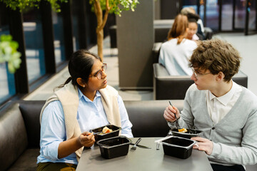 A White young man and an Indian young woman with glasses enjoying lunch together during their internship in a modern office space, engaging in conversation while working in a friendly environment.