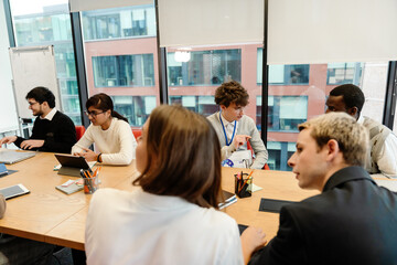 A diverse group of young professionals sits around a large table in a modern office. They engage in discussion and teamwork using tablets, notebooks, and documents during a collaborative meeting.