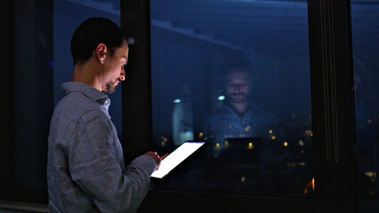 A man, engrossed in his work, stands by a window at night, illuminated by the glow of his tablet. The reflection of his focused demeanor is visible in the glass.
