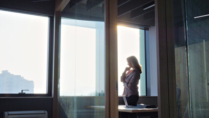 A woman stands near a window, gazing outside, symbolizing contemplation and focus in a modern office setting.