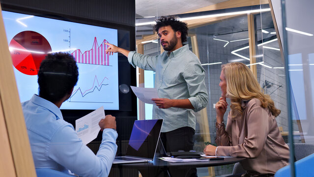 A man in a meeting explains a chart on a large screen to a group, highlighting the importance of effective communication and collaboration in a modern office setting.