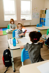 Obraz premium In a spacious classroom, a White boy holds colored pencils at his desk, with a Black girl sitting in front of him and a White girl behind him. Concept of school, classmates, and communication.