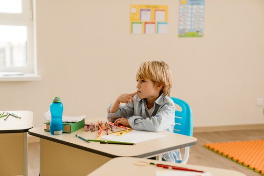 A White schoolboy with blonde hair sitting at a desk in a classroom, concentrating and biting a pencil while surrounded by school supplies, including colored pencils, a water bottle, and books.