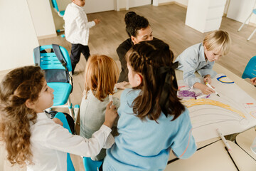 Multiracial school-aged boys and girls work together on a large group drawing with markers in a classroom. The children are engaged in a creative team activity.