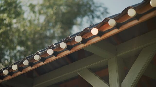 Charming shot of round bulbs along roof edge in bright daylight with blurred greenery background, highlighting tranquil outdoor setting and decorative lighting arrangement, creating cozy ambiance